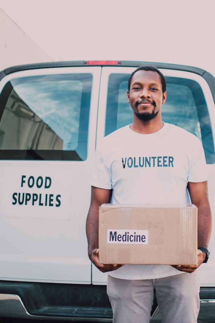 services-img Volunteer holding box of medicine in front of food supply van, showcasing community aid.