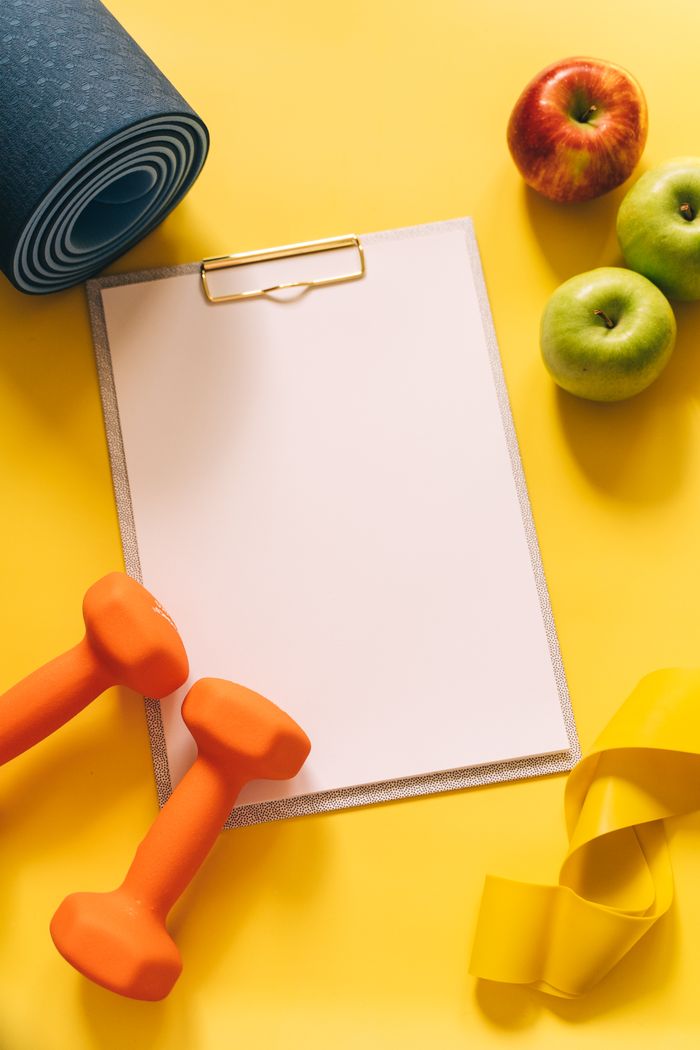 ours-journey Flat lay of gym equipment and clipboard for fitness planning on yellow background.